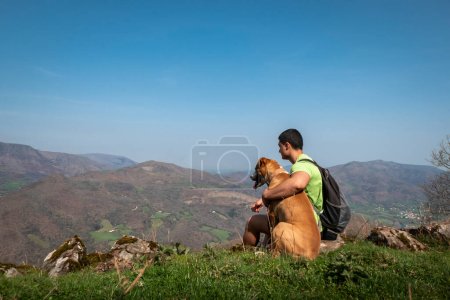 Joven turista sentado relajado con su perro y disfrutando de las montañas y pueblos al fondo de la montaña Jara en Iroulguy en el País Vasco