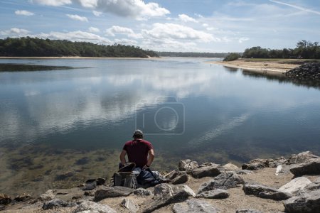 Téléchargez les photos : Pêcheur avec sa canne à pêche assise près de quelques rochers avec une petite île en arrière-plan couverte de forêt au Pays Basque, France - en image libre de droit