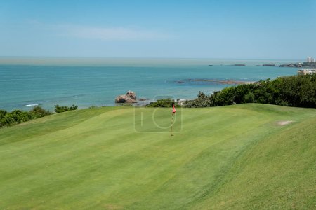 Téléchargez les photos : Terrain de golf avec un drapeau rouge au milieu et la mer en arrière-plan par une journée ensoleillée - en image libre de droit