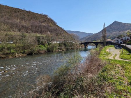 A small river meandering between mountains, flanked by a picturesque bridge and surrounding village on a sunny day