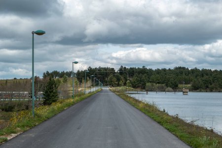 Road lined with electric lighting posts next to a water storage dam, surrounded by forest on a very cloudy day
