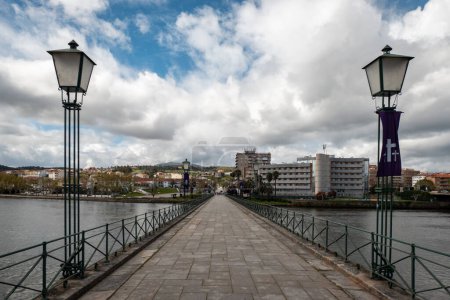 Pedestrian bridge over the Tua river in Mirandela with two old lamps as public lighting posts with part of the city in the background on a partly cloudy day in Portugal