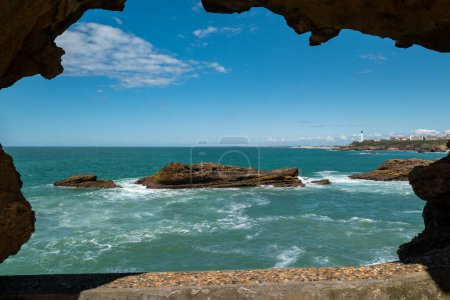 In the middle of a huge natural rock tunnel, some rocks in the sea with part of the city of Biarritz in the background on a sunny day