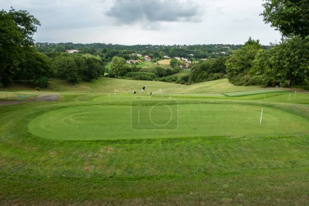 Golfplatz umgeben von grünen Wiesen und etwas Wald, im Hintergrund ein Golfer, der an einem sehr bewölkten Tag sein Können übt