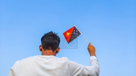 Boy holding Papua New Guinea flag against clear blue sky. Man hand waving Papuan flag view from back, copy space for text