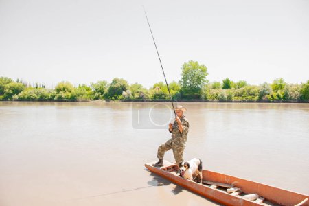 Photo for Focused man in camo clothes holding rod, checking hook, while sailing down river with dog on board. Side view of professional fisherman fishing, while standing on boat at daytime. Concept of fishing. - Royalty Free Image