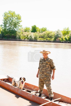 Photo for Concentrated male angler with faithful dog standing in old boat, going mooring after fishing in countryside. Front view of male fisherman enjoying trip on murky river. Concept of fishery, hobby. - Royalty Free Image