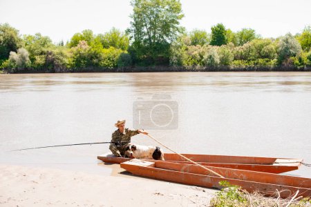Photo for Pensive male fisher with dog sailing from shore in shabby boat in sunny day. Front view of man with stick pushing off from bank, going fishing, with calm river on background. Concept of fishery. - Royalty Free Image