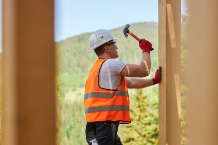 Photo for Side view of male builder wearing yellow vest and helmet working outdoors, building wooden house. Man holding hammer, raising, beating nail in wooden board. Concept of building. - Royalty Free Image