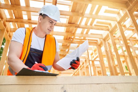 Photo for Side view of male architect, engineer, builder standing, constructing building, planning project. Young man wearing yellow vest and helmet writing, looking down. Concept of building. - Royalty Free Image