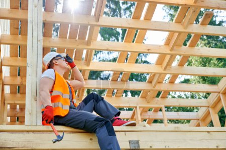 Photo for Side view of worker wearing yellow vest, white helmet, glasses, building wooden house in forest. Professional man sitting, holding hammer, having rest. Concept of building. - Royalty Free Image