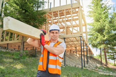 Photo for Side view of builder, worker wearing uniform and helmet standing, holding wooden gilder, board, carrying. Man wearing vest and helmet looking aside, smiling. Concept of building. - Royalty Free Image