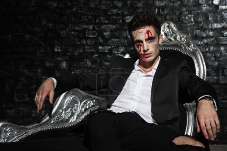 Porträt eines gutaussehenden jungen Mannes, der im Anzug auf einer Couch sitzt, sein Gesicht blutig geschminkt hat und ernst aussieht