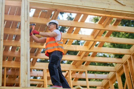 Photo for Side view of strong builder standing on wooden building, putting gilder, holding hammer, beating. Man wearing uniform and helmet building wooden house. Concept of building. - Royalty Free Image