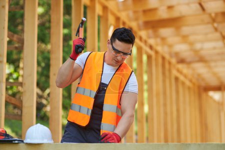 Photo for Front view of builder wearing vest and glasses, putting off helmet, holding hammer, beating nail. Man building wooden house in forest, putting gilder. Concept of building. - Royalty Free Image