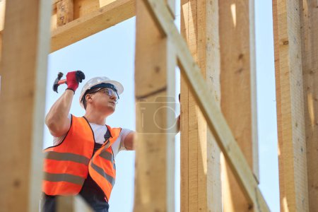 Photo for Side view of builder wearing helmet and uniform, standing, holding hammer, beating nail in wooden board. Man working outdoors, building wooden house. Concept of building. - Royalty Free Image