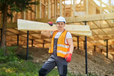 Photo for Front view of man, worker wearing uniform and helmet, standing, holding wooden boards, gilders. Builder constructing wooden house, looking at camera. Concept of building. - Royalty Free Image