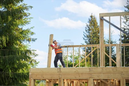 Photo for Side view of strong man, worker, craftsman building, constructing wooden house in forest in summer. Builder wearing uniform and helmet, holding gilder. Concept of building. - Royalty Free Image