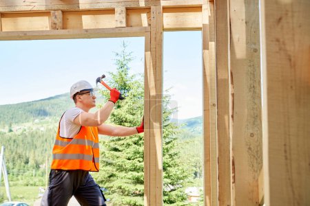 Photo for Side view of professional worker, builder standing, holding hammer and wooden gilder, beating. Man wearing vest and helmet, building wooden house in forest. Concept of building. - Royalty Free Image