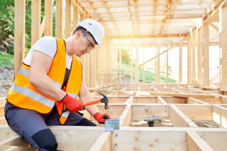 Photo for Side view of builder wearing vest an helmet, sitting, holding hammer, beating nail in wooden decking. Young man working outdoors, constructing house. Concept of building. - Royalty Free Image
