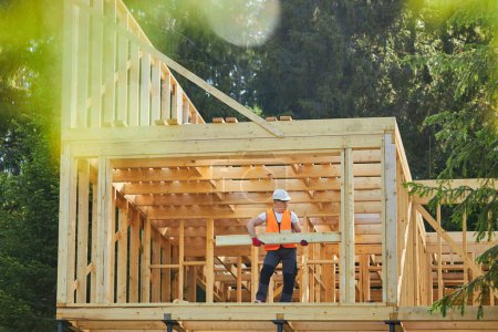 Photo for Front view of man, builder, worker building wooden house in forest, in sunny weather, in summer. Male wearing helmet and uniform holding, putting gilder. Concept of building. - Royalty Free Image