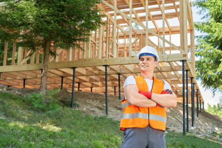 Photo for Front view of young man, builder wearing uniform and helmet, standing with crossed hands, admiring forest. Engineer, architect looking forward, smiling. Concept of building. - Royalty Free Image