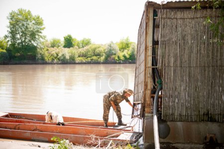 Photo for Fisherman in camouflage outfit untying rowing boat from jetty, while going fishing with dog along river. Side view of busy man untangling metal chain of skiff at dock, sailing away. Concept of rest. - Royalty Free Image