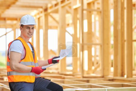 Photo for Front view of young architect, builder, engineer constructing wooden house in forest in summer. Man wearing uniform and helmet, sitting, holding paper. Concept of building. - Royalty Free Image