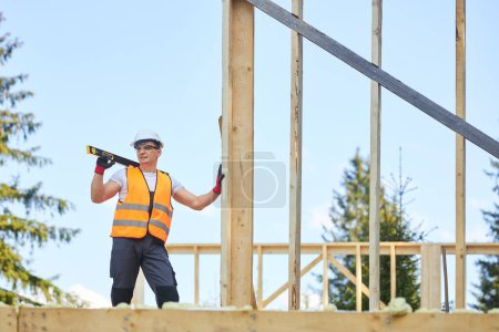 Photo for Side view of man wearing helmet and uniform standing on wooden concstrction, holding measuring tool. Builder, worker building house in forest in summer. Concept of building. - Royalty Free Image