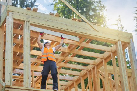 Photo for Front view of builder wearing uniforn, vest and helmet, holding, raising wooden gilder, board. Man, worker building wooden house in forest in summer. Concept of building. - Royalty Free Image