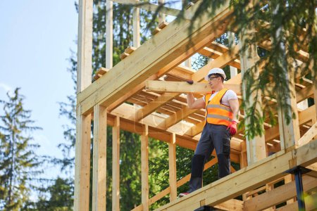 Photo for Side view of strong male builder, holding, raising, carrying wooden board, gilder, looking forward. Mna wearing uniform and helmet constructing wooden house. Concept of building. - Royalty Free Image