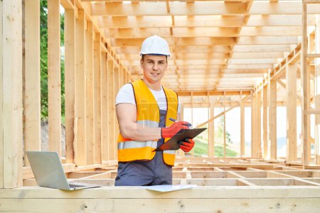 Photo for Front view of young architect, builder, engineer standing in wooden building, looking at camera. Man wearing vest and helmet constructing building, planning. Concept of building. - Royalty Free Image