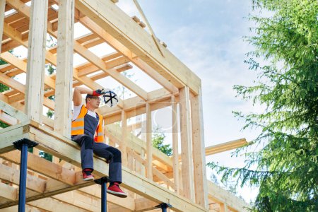 Photo for Side view of male worker, builder sitting high, having rest, looking forward, putting off helmet. Professional craftsman building wooden house in forest. Concept of building. - Royalty Free Image