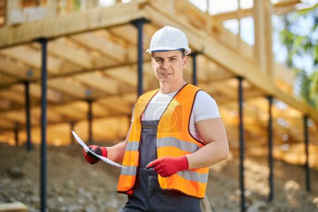 Photo for Front view of young man wearing yellow vest, uniform and white helmet, standing, building house. Builder, engineer planning project, looking at camera. Concept of building. - Royalty Free Image