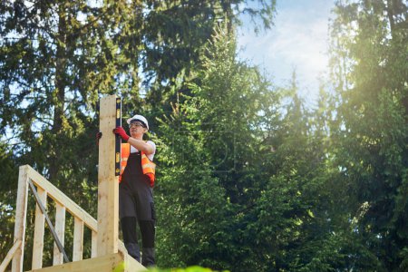 Photo for Front view of young builder wearing uniform, helmet and glasses building house in forest in summer. Worker standing, putting wooden gilder, measuring. Concept of building. - Royalty Free Image