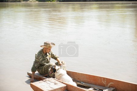 Photo for Concentrated fisherman wetting hand in muddy water, while sitting in boat with dog. Side view of tired male angler in camo having rest, freshing up during fishery. Pastime, fishing concept. - Royalty Free Image
