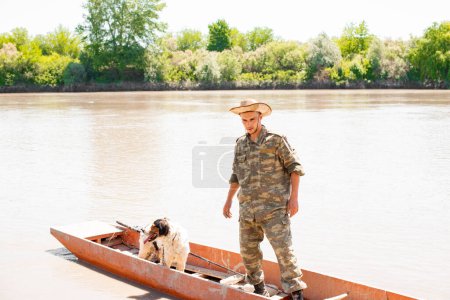 Photo for Focused young angler standing in old red skiff, thinking, going mooring to pier after fishery at day. Front view of male fisher with dog enjoying boat trip on calm river. Concept of fishery, hobby. - Royalty Free Image