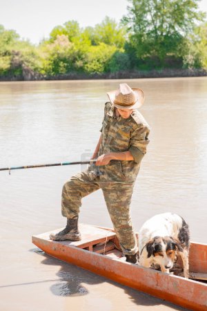 Photo for Fisherman in camo clothes holding fishing rod, while standing on stern of boat and fishing. Front view of professional angler looking down, while floating with dog on skiff at day. Concept of fishing. - Royalty Free Image