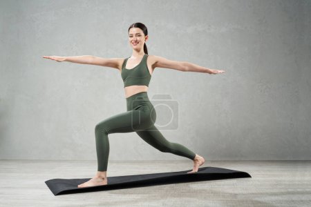 Photo for Happy female with ponytail staying in Virabhadrasana II against grey wall. Side view of flexible fit woman smiling to camera, while practicing warrior II pose in yoga class studio. Lifestyle concept. - Royalty Free Image