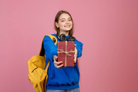 Front view of stylish schoolgirl standing, holding gift box, showing. Pretty brunette girl wearing casual clothes, loking at camera, smiling. Isolated on pink studio background.