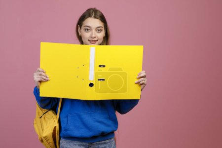 Front view of pretty schoolgirl standing, holding open folder, showing. Beautiful young female brunette holding yellow rucksack, looking at camera, smiling. Concept of youth.