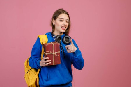 Front view of young student holding gift box, showing super and tongue. Pretty young brunette female standing, looking at camera, winking. Isolated on pink studio background.