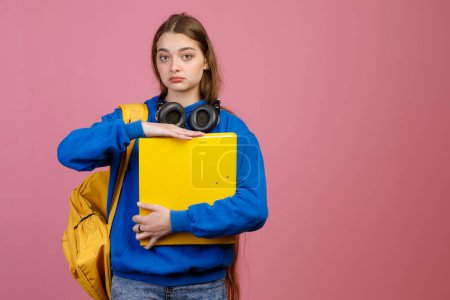 Front view of brunette schoolgirl standing, holding folder by hands. Pretty young female looking at camera, with blue khudi and jeans, earphones. Isolated on pink background.