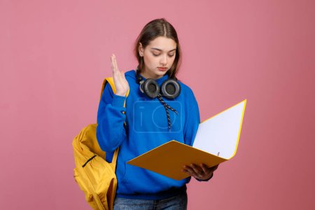 Front view of pretty schoolgirl standing, reading, raising hand, answering. Beautiful young female with yellow rucksack and earphones studying. Concept of youth and school life.