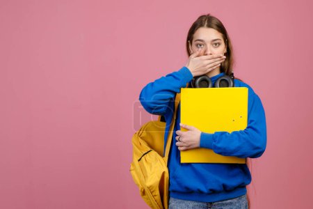 Front view of shocked, surprised schoolgirl standing, closing mouth by hand. Pretty young female student holding yellow rucksack and folder. Isolated on pink studio background.