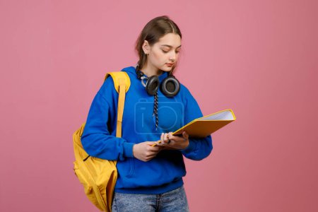 front view of schoolgirl with earphones standing, holding yellow folder, reading. Pretty brunette female looking down, wearing blue khudi and jeans. Isolated on pink background.