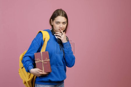 Front view of pretty brunette schoolgirl touching lips, remembering. Young student standing, holding bunch of old books, looking at camera. Concept of education and youth.