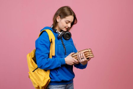 Side view of teenage girl standing, looking down. Pretty brunette schoolgirl with earphones holding bunch of old books and yellow rucksack. Isolated on pink studio background.