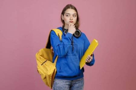 Front view of young student standing, touching chin, thinking. Pretty schoolgirl wearing blue khudi and jeans looking aside, holding yellow folder. Concept of education and youth.