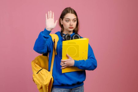 Front view of teenage femal standing, holding yellow backpack and folder. Pretty young schoolgirl looking forward, raising hand, greeting. Isolated on pink studio background.